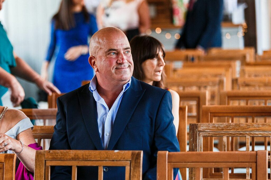 A bald man in a navy blue suit sits on a wooden chair at what appears to be a formal event or gathering. Other attendees wearing various formal clothes are seen in the background, engaging in conversation and enjoying the event. Image by Documentary Wedding Photographer Lewis Bishop Photography.