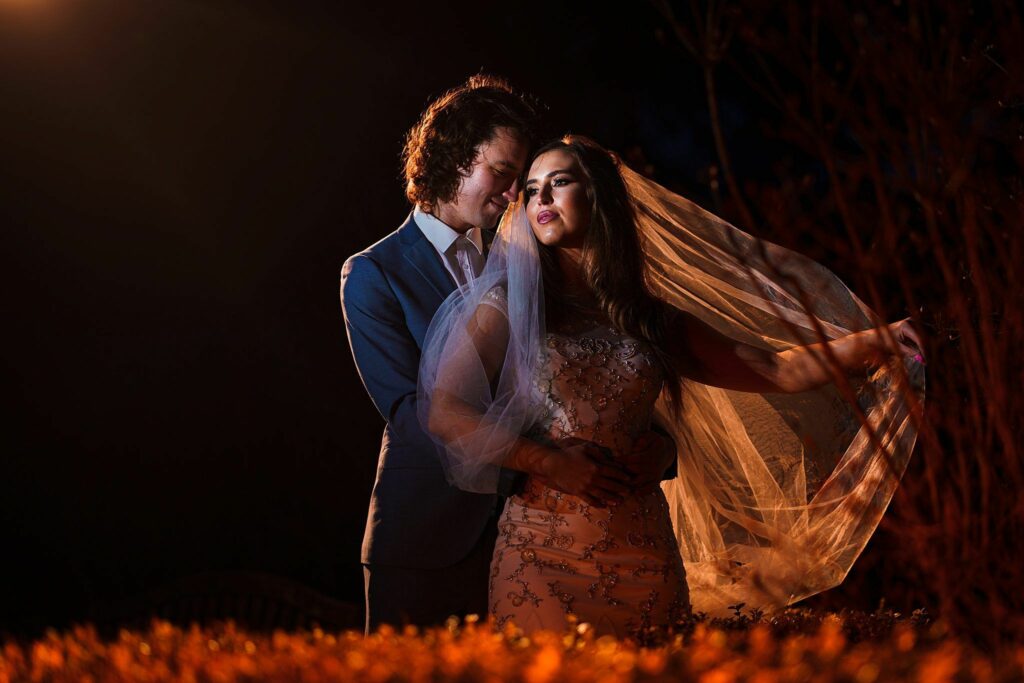 A couple is standing closely together at night, with the man embracing the woman from behind. The woman is wearing a veil and a decorated dress, while the man is dressed in a dark suit. Warm lighting highlights them against a dark background and foliage in the foreground. Image by Documentary Wedding Photographer Lewis Bishop Photography.