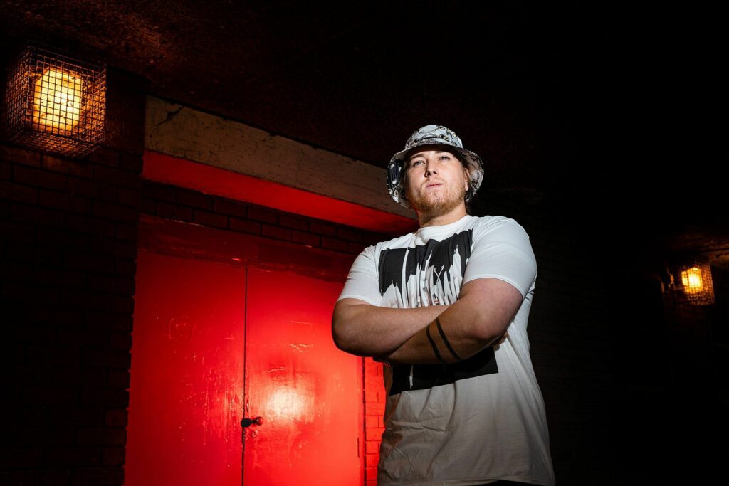 A person wearing a white t-shirt and a patterned bucket hat stands with arms folded in front of a red door, illuminated by overhead lights. The background is dimly lit, and the person appears relaxed and confident. Image by Documentary Wedding Photographer Lewis Bishop Photography.