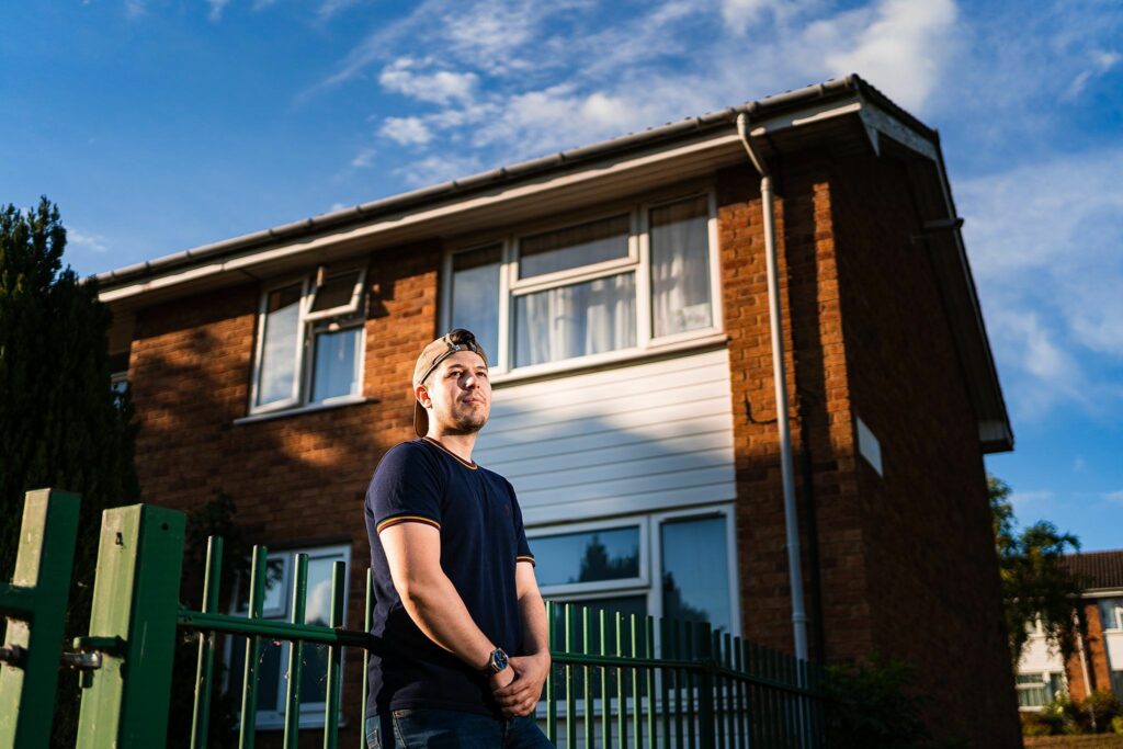 A person in a navy blue shirt and beige hat stands outside a brick house with white paneling, looking off into the distance. The sky behind is blue with some clouds. A green fence is in the foreground. Image by Documentary Wedding Photographer Lewis Bishop Photography.