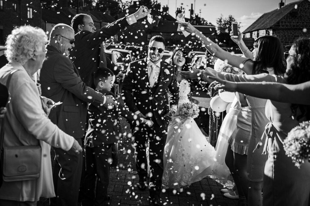 A newlywed couple walks down a path as friends and family shower them with flower petals. The groom, wearing sunglasses, smiles widely. The bride holds a bouquet and grins joyfully. Guests around them celebrate, some holding up their phones to capture the moment. Image by Documentary Wedding Photographer Lewis Bishop Photography.
