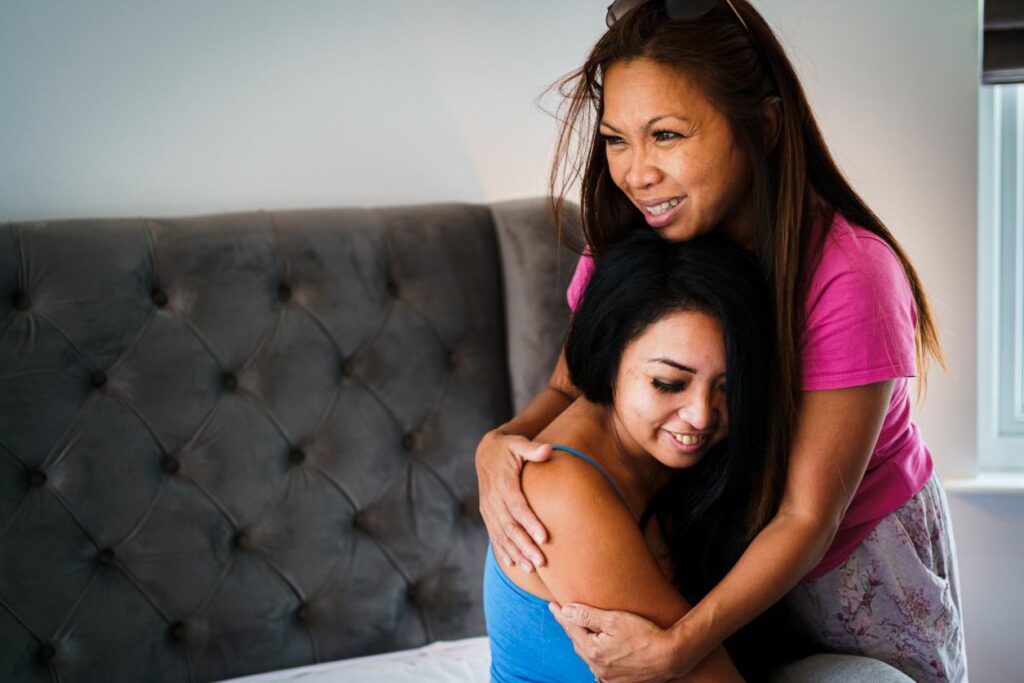 Two women are embracing each other warmly. One is seated on a bed, smiling with her eyes closed, while the other, standing beside her, gently hugs her with a comforting expression. They are in a cozy, well-lit room with a tufted headboard visible in the background. Image by Documentary Wedding Photographer Lewis Bishop Photography.