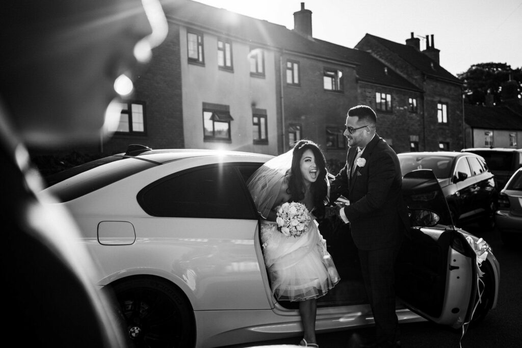 A joyful bride, holding a bouquet, steps out of a white car with the assistance of a suited man, possibly the groom. The scene is set on a sunny day with light shining through, casting a bright, cheerful atmosphere. In the background are brick houses and a few parked cars. Image by Documentary Wedding Photographer Lewis Bishop Photography.