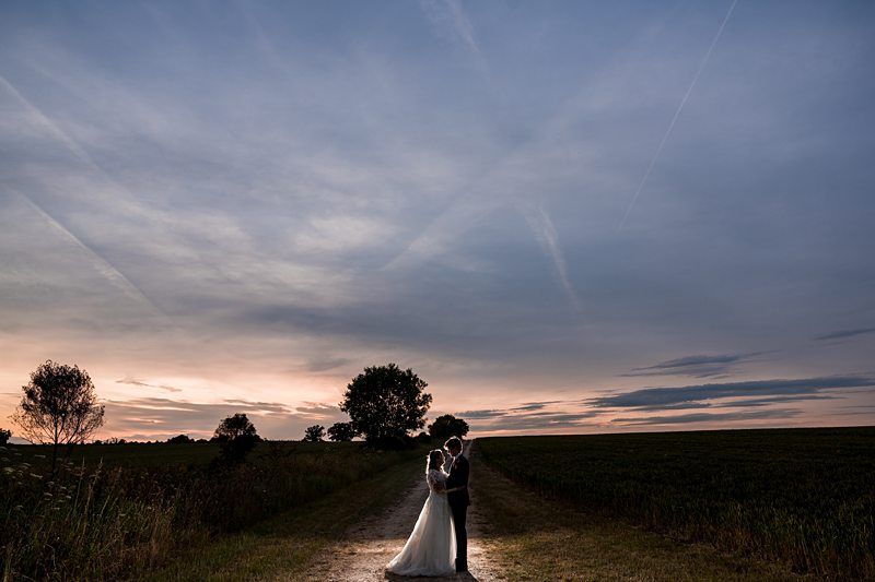 A couple wearing wedding attire embraces on a rural path at sunset, with a tree and grassy fields in the background under a partially cloudy sky. Image by Documentary Wedding Photographer Lewis Bishop Photography.