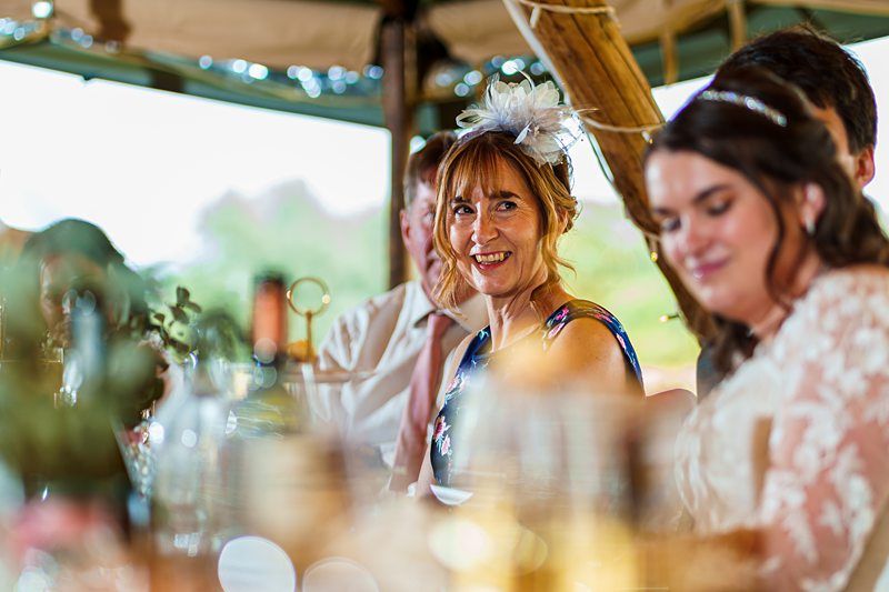 A woman wearing a floral dress and fascinator smiles while sitting at a table, with other people in the background at a social event. Image by Documentary Wedding Photographer Lewis Bishop Photography.