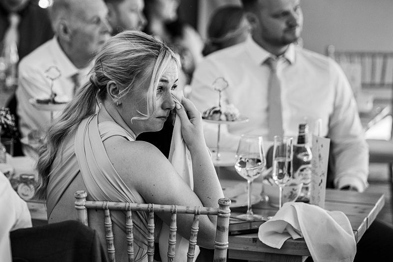 A woman wipes her tears with a tissue while seated at a table during an event. Other attendees are visible in the background, seated, and appear to be engaged. Image by Documentary Wedding Photographer Lewis Bishop Photography.