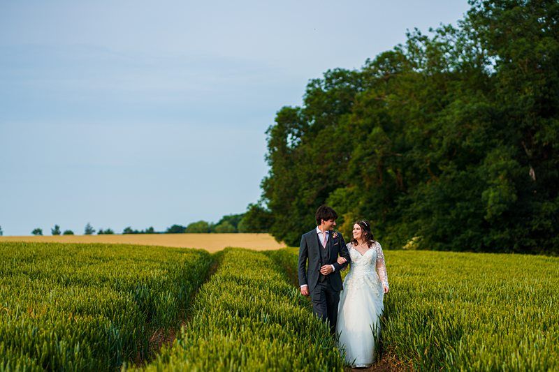 A couple in wedding attire walks hand-in-hand through a lush green field, with trees in the background under a clear sky. Image by Documentary Wedding Photographer Lewis Bishop Photography.