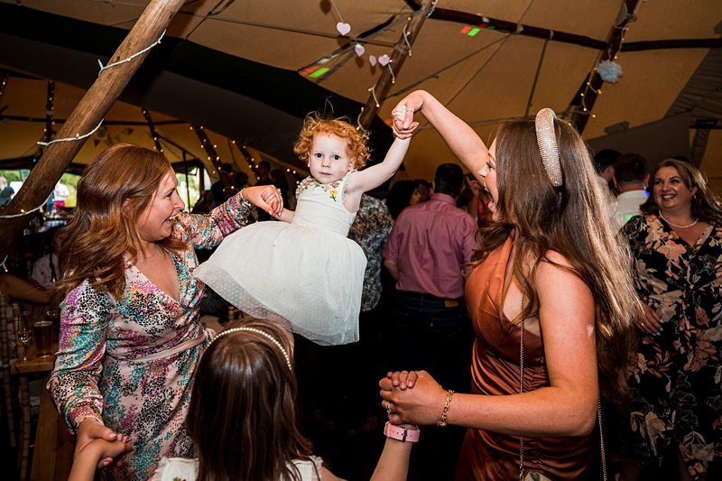 A girl in a white dress is lifted by two women holding her hands, with people dancing and smiling around them at an indoor event. Image by Documentary Wedding Photographer Lewis Bishop Photography.