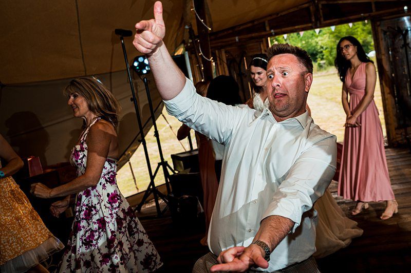 A man in a white shirt exuberantly dances, pointing with both hands, while others dance in the background inside a tent-like structure. Image by Documentary Wedding Photographer Lewis Bishop Photography.