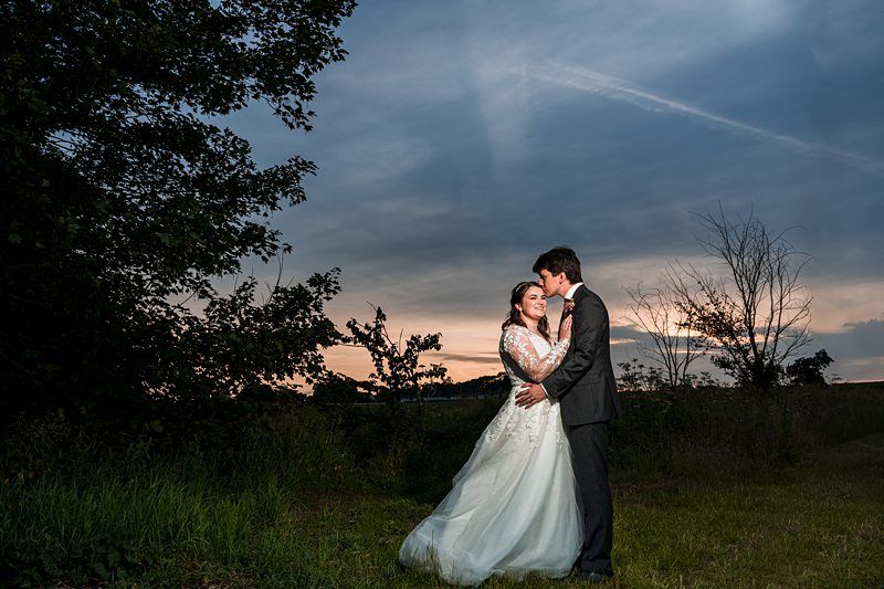 A bride and groom stand close together in an outdoor setting at dusk. The bride is in a white gown, and the groom is in a dark suit. Trees and a colorful sky are visible in the background. Image by Documentary Wedding Photographer Lewis Bishop Photography.