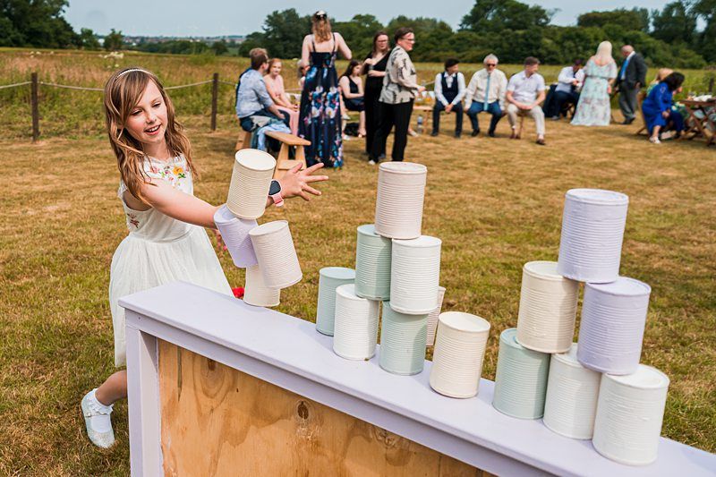 A young girl is playing a game of tin can knockdown at an outdoor event, with several people seated and standing in the background on a grassy field. Image by Documentary Wedding Photographer Lewis Bishop Photography.