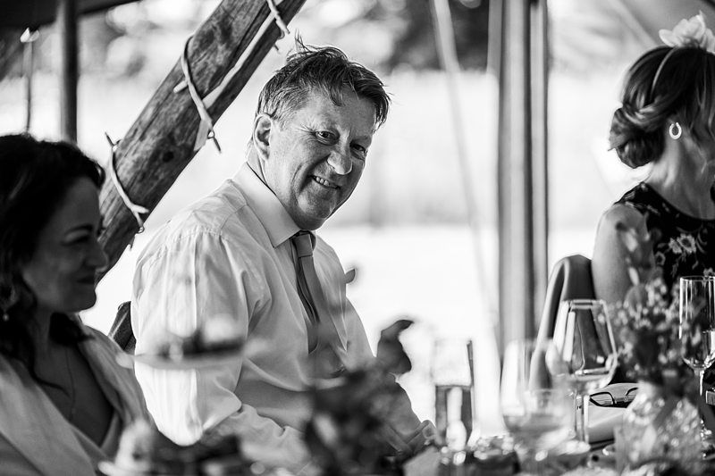 A man in a tie smiles while sitting at a table, surrounded by other people. The setting appears to be an event or gathering, with food and drinks on the table. The image is in black and white. Image by Documentary Wedding Photographer Lewis Bishop Photography.