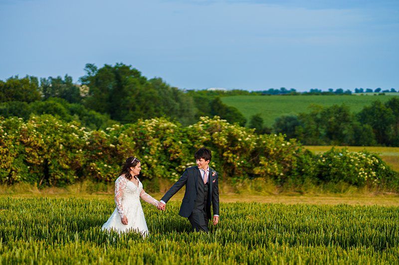 A bride in a white dress and a groom in a dark suit walk hand in hand through a field of tall grass with trees and a clear sky in the background. Image by Documentary Wedding Photographer Lewis Bishop Photography.