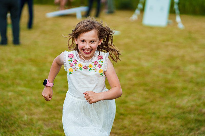 A young girl in a white dress with floral embroidery runs on a grassy field, smiling. She is wearing a pink wristwatch. Other people and objects are blurred in the background. Image by Documentary Wedding Photographer Lewis Bishop Photography.