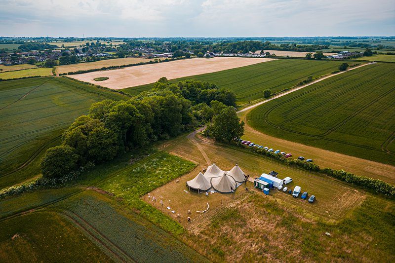 Aerial view of a rural area with vast green fields, a group of tents set up near trees, and several parked cars nearby. Image by Documentary Wedding Photographer Lewis Bishop Photography.