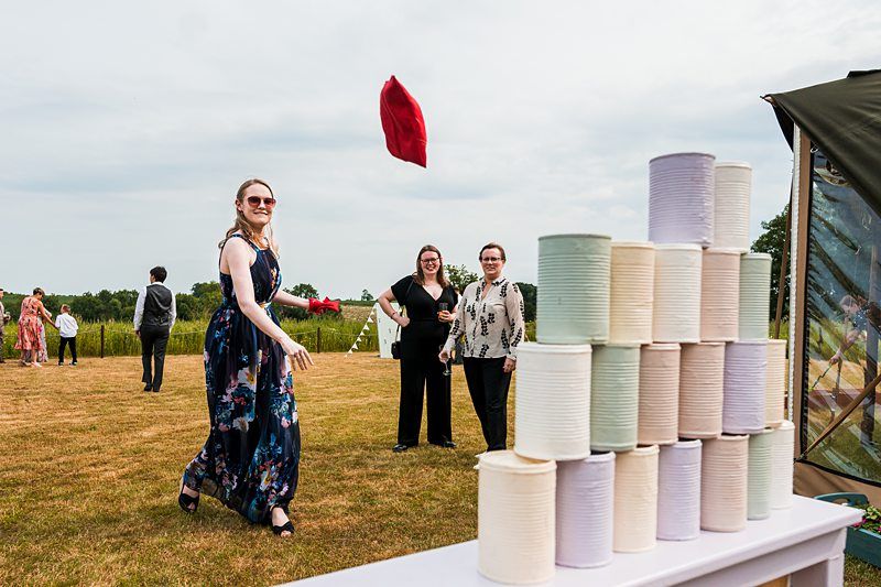 A woman in a floral dress is tossing a red beanbag toward a stack of tin cans while two other women watch. The scene takes place outdoors on a grassy area. Image by Documentary Wedding Photographer Lewis Bishop Photography.