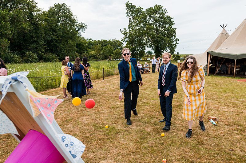 People play an outdoor game involving throwing balls at a target in a grassy area. They are casually dressed and appear to be at a social event or gathering, with a tent visible in the background. Image by Documentary Wedding Photographer Lewis Bishop Photography.