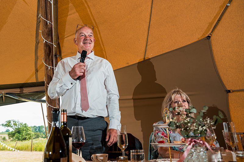 A man wearing a white shirt and pink tie stands holding a microphone under a tent. A woman sits at a decorated table nearby. Various drinks and flowers are on the table. Image by Documentary Wedding Photographer Lewis Bishop Photography.