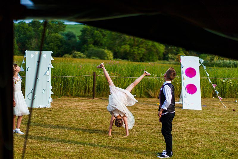 A young girl performs a cartwheel on a grassy field while two children watch, with cardboard targets and a green landscape in the background. Image by Documentary Wedding Photographer Lewis Bishop Photography.