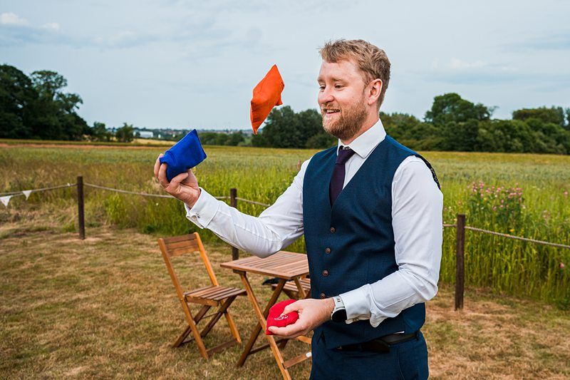 A man with a beard and a blue vest juggles three colorful beanbags in an outdoor setting with chairs and fields in the background. Image by Documentary Wedding Photographer Lewis Bishop Photography.
