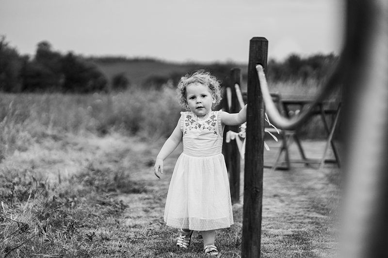 A young girl in a white dress with floral details walks along a rope fence in a grassy outdoor area, holding onto a post. The background features trees and open fields. Black and white photo. Image by Documentary Wedding Photographer Lewis Bishop Photography.