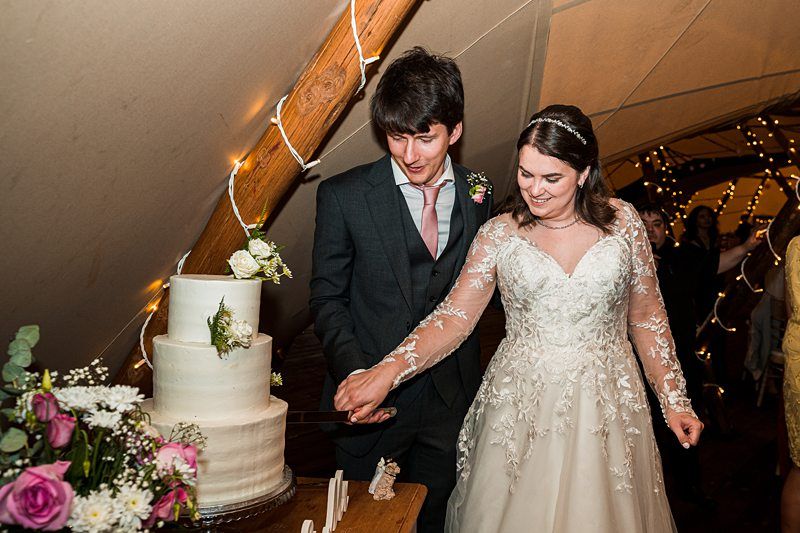 A bride and groom cut a three-tiered wedding cake together under a tent decorated with string lights. The bride wears a white lace gown, and the groom is in a dark suit with a pink tie. Image by Documentary Wedding Photographer Lewis Bishop Photography.