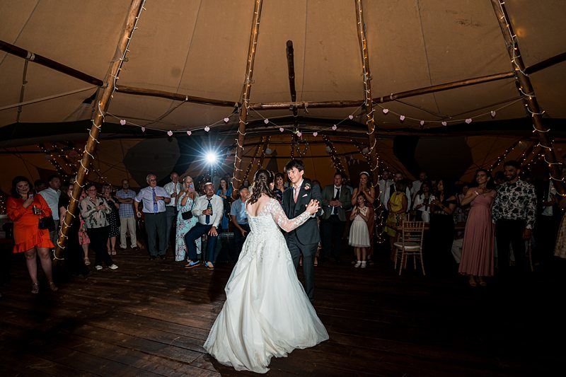 A bride and groom, dressed in wedding attire, dance under a tent with guests watching and string lights overhead. Image by Documentary Wedding Photographer Lewis Bishop Photography.