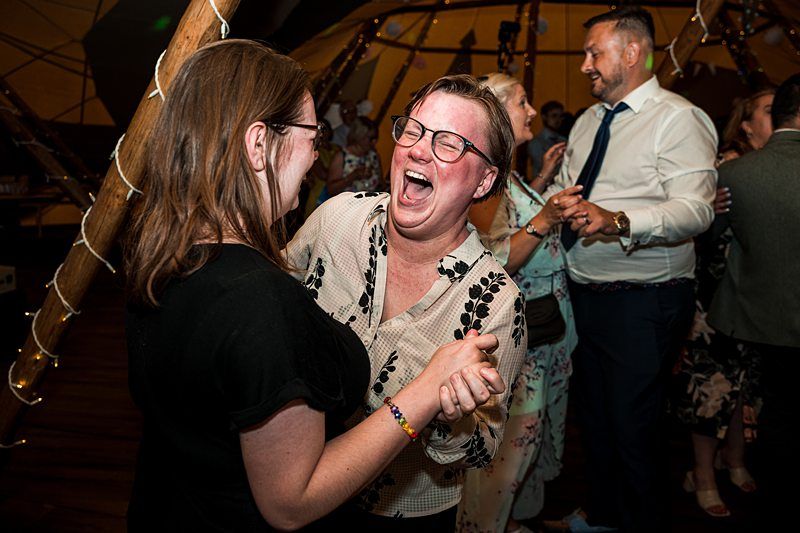 Two people laughing and dancing closely at a lively indoor event, with other smiling attendees in the background. Image by Documentary Wedding Photographer Lewis Bishop Photography.