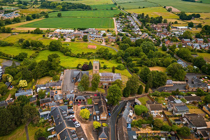 Aerial view of a rural village featuring a prominent stone church with a bell tower, surrounded by residential buildings, trees, and green fields in the background. Image by Documentary Wedding Photographer Lewis Bishop Photography.
