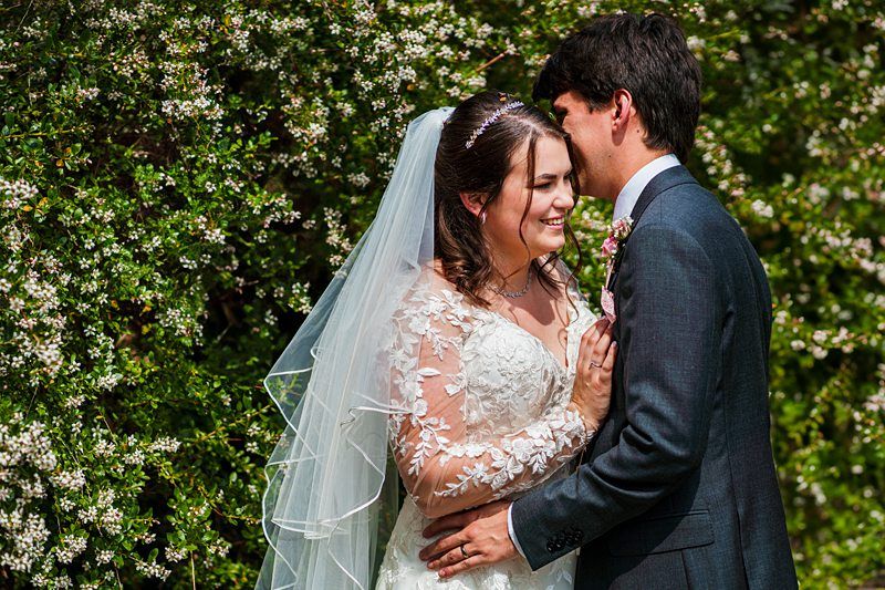 A bride and groom stand closely together, smiling and embracing, with a backdrop of green foliage and white flowers. The bride wears a white dress and veil, while the groom wears a dark suit. Image by Documentary Wedding Photographer Lewis Bishop Photography.