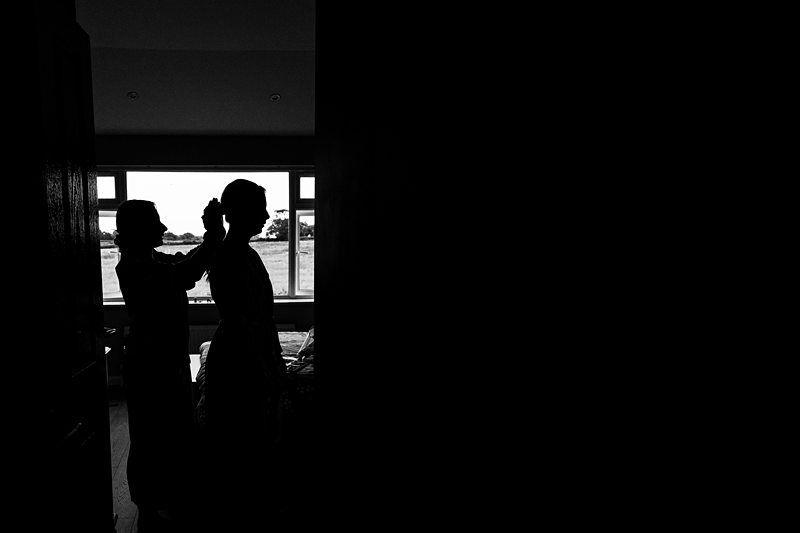 Silhouette of two people indoors, one adjusting the other's attire, with a bright window in the background. Image by Documentary Wedding Photographer Lewis Bishop Photography.
