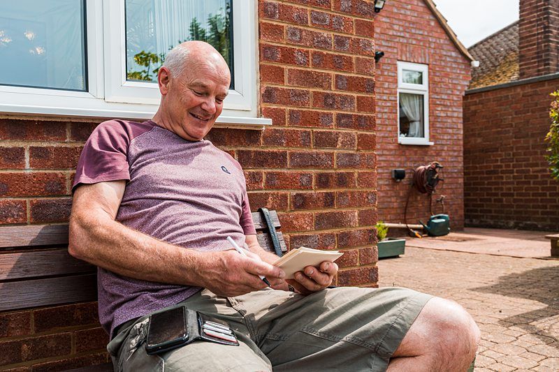 A man with short gray hair sits on a bench outside a brick house, smiling while writing on a notepad. He wears a purple and gray t-shirt and khaki shorts, with a wallet and phone on his lap. Image by Documentary Wedding Photographer Lewis Bishop Photography.