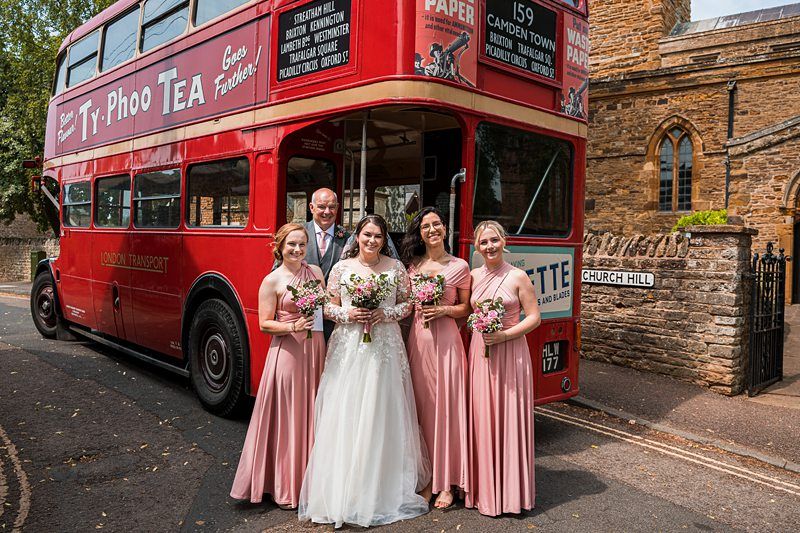 A bride in a white gown stands with four bridesmaids in pink dresses holding bouquets, in front of a red double-decker bus on a street near a church. Image by Documentary Wedding Photographer Lewis Bishop Photography.