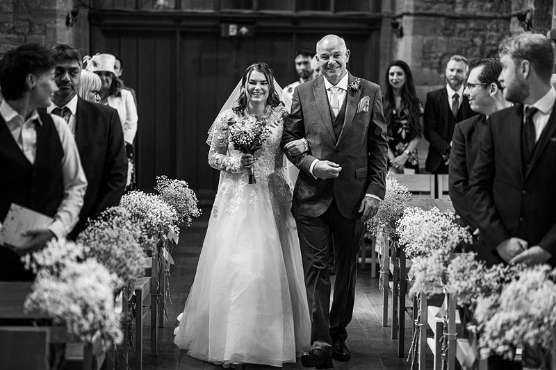 A bride in a white dress holding a bouquet walks down the aisle with a man in a suit, surrounded by seated guests in a decorated church. Image by Documentary Wedding Photographer Lewis Bishop Photography.