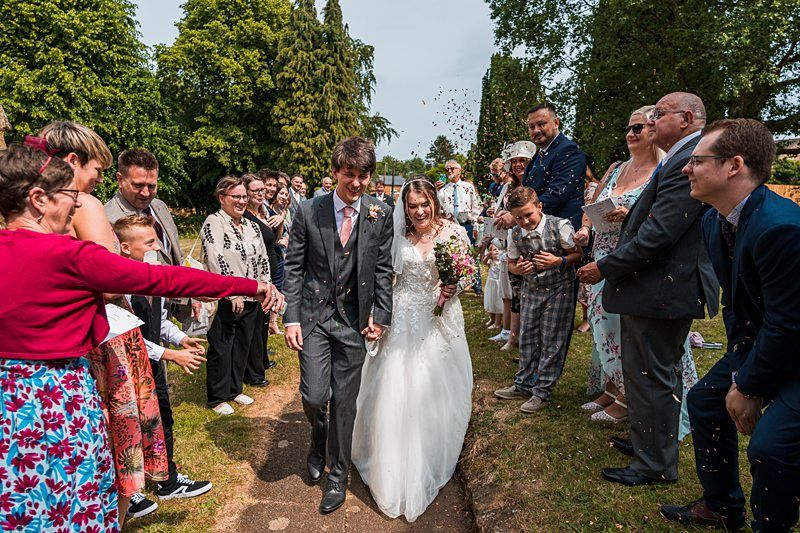 A newlywed couple walks down an outdoor path, holding hands and smiling, surrounded by guests who are throwing confetti and celebrating. Image by Documentary Wedding Photographer Lewis Bishop Photography.