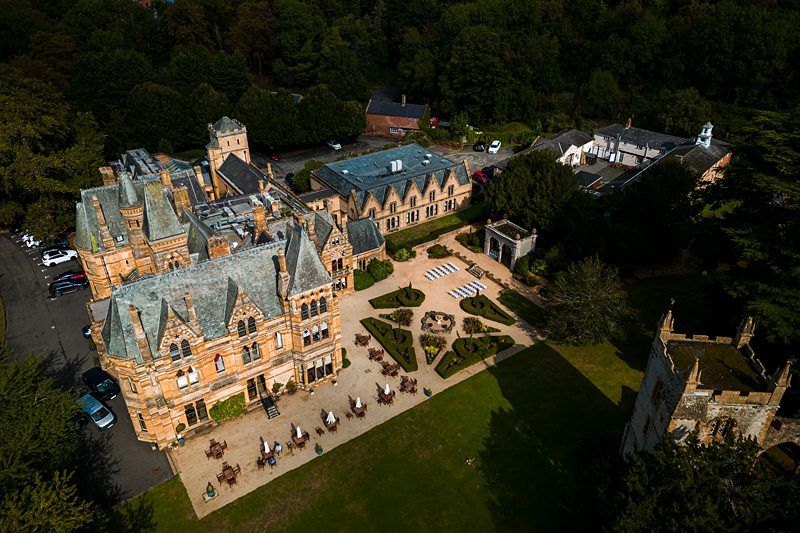 Aerial view of a large, historic mansion with elaborate architecture and multiple gabled roofs, surrounded by landscaped gardens and trees. The courtyard area features outdoor seating and geometric garden designs. Several cars are parked nearby on the left. Image by Documentary Wedding Photographer Lewis Bishop Photography.