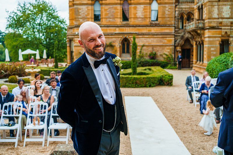A smiling groom in a black velvet tuxedo with a bow tie stands at the end of an outdoor aisle, awaiting the bride. Guests are seated along the aisle in white chairs. The wedding venue is a grand, historic building with intricate architecture and green gardens. Image by Documentary Wedding Photographer Lewis Bishop Photography.