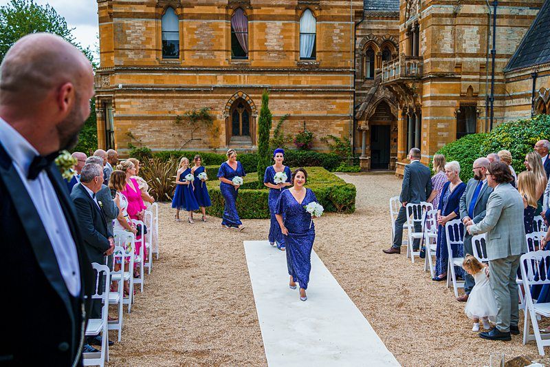 A formal outdoor wedding with a historic building in the background. Bridesmaids in matching blue dresses walk down a white aisle. Guests in elegant attire are seated on both sides, and a man in a suit is visible in the foreground, turning to watch the procession. Image by Documentary Wedding Photographer Lewis Bishop Photography.