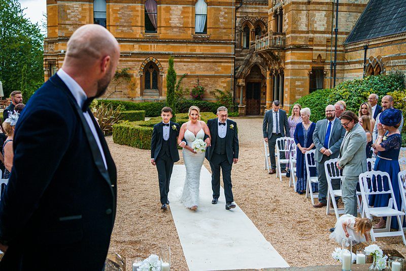 A bride in a white dress, holding a bouquet, walks down an outdoor aisle with two men. Guests in formal attire stand and watch on either side of the aisle. The setting appears to be a historic stone building. Another man, possibly the groom, watches from the foreground. Image by Documentary Wedding Photographer Lewis Bishop Photography.