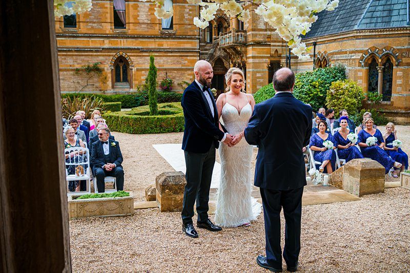 A couple stands hand in hand before an officiant during an outdoor wedding ceremony. The bride wears a white gown, and the groom is dressed in a dark suit. Guests, seated on either side, watch attentively. The setting includes a historic building and lush greenery. Image by Documentary Wedding Photographer Lewis Bishop Photography.