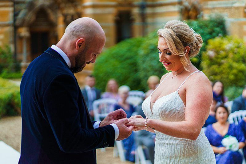A couple stand outdoors in a garden, facing each other during a wedding ceremony. The woman, wearing a white dress, is smiling while the man, in a dark suit, places a ring on her finger. Guests are seated in the background, watching the ceremony. Image by Documentary Wedding Photographer Lewis Bishop Photography.