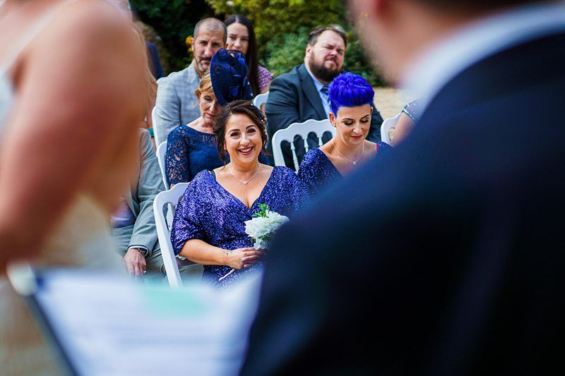 Several people are seated outdoors at a wedding ceremony. In the foreground, blurred, someone is holding papers. A smiling guest in a blue dress and fascinator holds a bouquet, while another guest with bright blue hair sits next to her, looking down. Image by Documentary Wedding Photographer Lewis Bishop Photography.