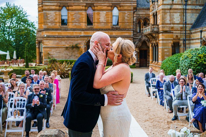 A newlywed couple shares a kiss outdoors in front of an ornate, historic building. The bride is in a white dress, and the groom is in a dark suit. Guests seated on either side of the aisle watch and applaud, while trees and a garden surround the venue. Image by Documentary Wedding Photographer Lewis Bishop Photography.