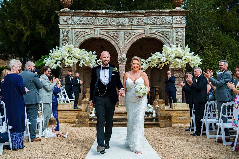 A couple dressed in wedding attire walks down an outdoor aisle while guests on both sides clap and cheer. The groom wears a black tuxedo, and the bride wears a white gown, holding a bouquet. The aisle is lined with chairs and the backdrop features a decorative arch. Image by Documentary Wedding Photographer Lewis Bishop Photography.