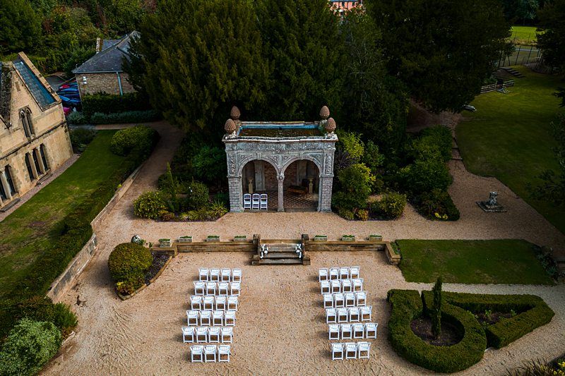 Aerial view of an outdoor wedding setup with white chairs arranged in rows facing a small ornate archway. The archway is situated in a garden area between two large buildings, surrounded by lush greenery and pathways. Image by Documentary Wedding Photographer Lewis Bishop Photography.