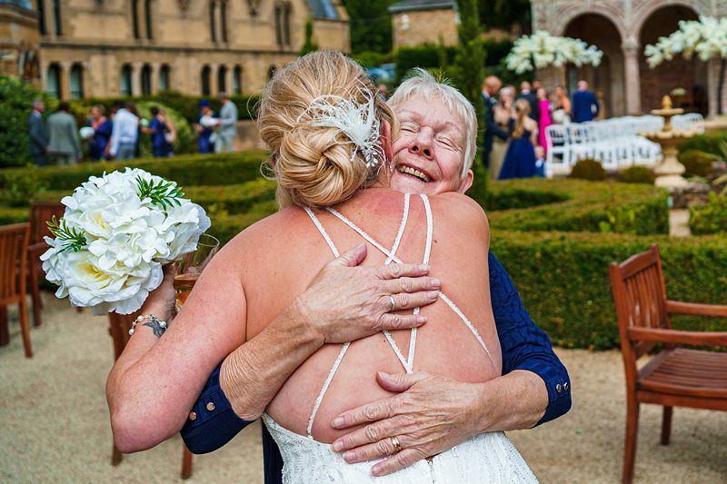 A bride in a white dress, holding a bouquet of white flowers, hugs an elderly woman in a navy dress outside at a wedding venue. They are surrounded by garden hedges and guests in the background. Both appear to be smiling and joyful. Image by Documentary Wedding Photographer Lewis Bishop Photography.