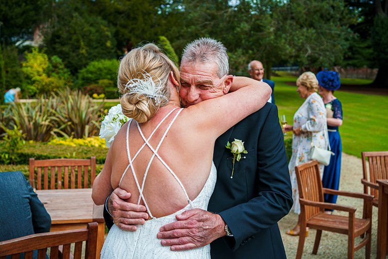 A bride in a white dress with crisscross straps hugs an older man in a suit and boutonnière outside in a garden setting. Several well-dressed guests can be seen in the background, enjoying the event in a lush, green environment. Image by Documentary Wedding Photographer Lewis Bishop Photography.