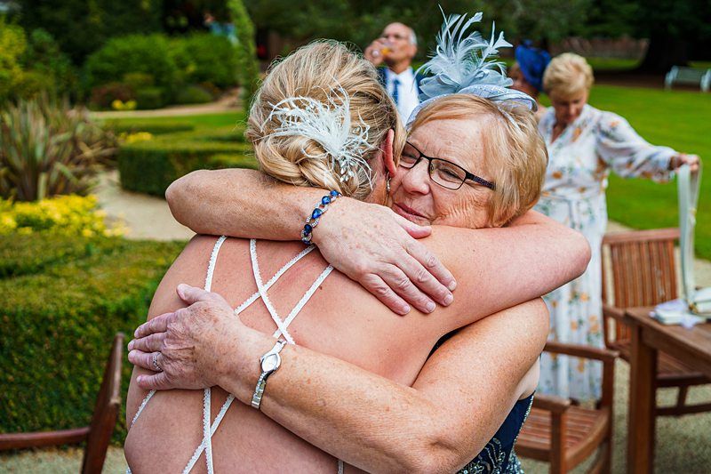 An older woman with glasses hugs a younger woman wearing a strappy dress, both with feathered hair accessories. They appear to be at an outdoor garden event with manicured bushes and other people in the background. Image by Documentary Wedding Photographer Lewis Bishop Photography.