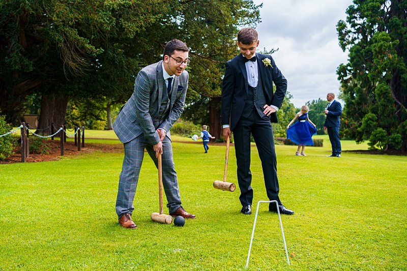 Two men in suits play croquet on a grassy lawn. One, in a grey suit, is leaning down and preparing to hit a croquet ball with a mallet, while the other, in a black suit, watches. People in formal attire and a child are visible in the background. Image by Documentary Wedding Photographer Lewis Bishop Photography.