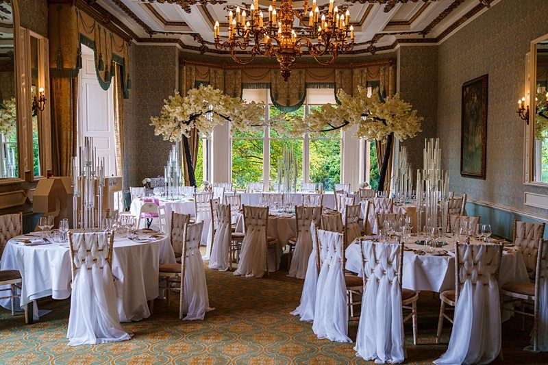 A beautifully decorated banquet hall with round tables draped in white linens and matching chairs adorned with white fabric bows. Tall floral centerpieces and elegant candle arrangements enhance the refined atmosphere. Large windows offer a view of lush greenery. Image by Documentary Wedding Photographer Lewis Bishop Photography.